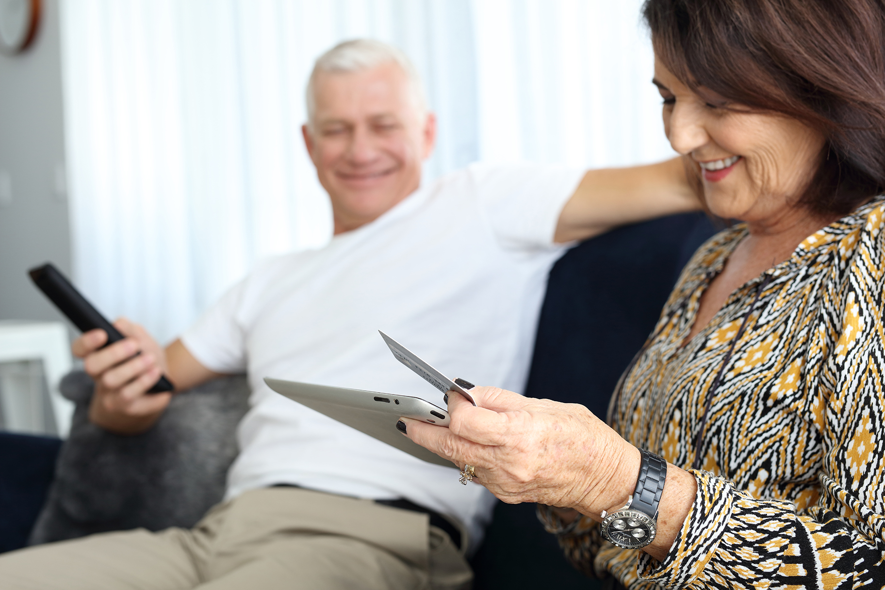 Man holding TV remote woman looking at tablet with credit card on couch