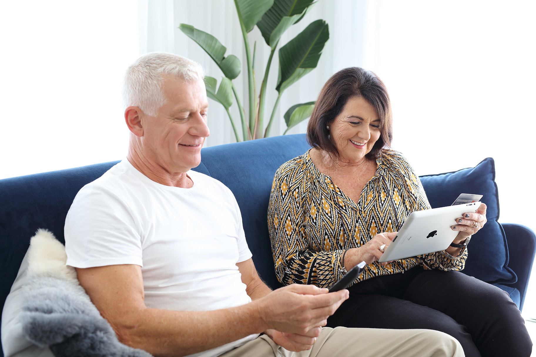 Man holding TV remote woman looking at tablet with credit card on couch