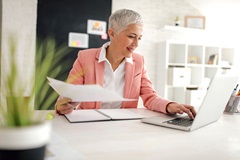 Business woman in office looking at laptop holding paperwork