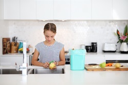 Girl peeling potato at kitchen sink with bin on bench beside her